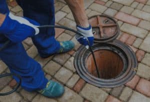 A Pipeworks Plumbing and Construction technician in blue workwear and gloves uses a diagnostic tool to inspect an open outdoor manhole for basement floor drain backup on a brick-paved surface.