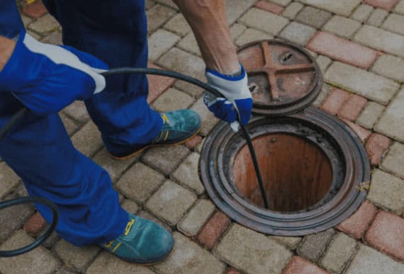 A Pipeworks Plumbing and Construction technician in blue workwear and gloves uses a diagnostic tool to inspect an open outdoor manhole for basement floor drain backup on a brick-paved surface.