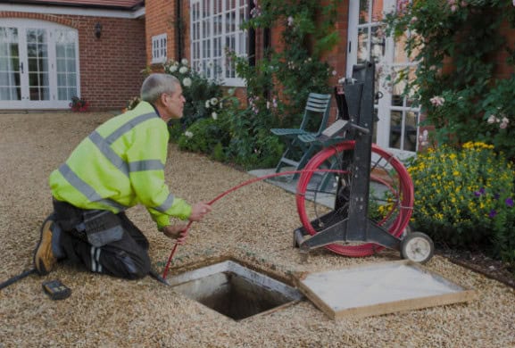 A Pipeworks Plumbing and Construction technician in a high-visibility jacket kneels on gravel, expertly installing piping by guiding a red cable into an open ground utility hole outside a brick house with landscaped gardens and flowers.