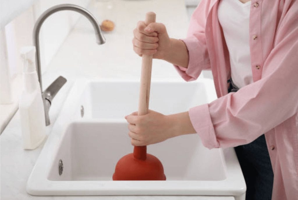 Pipeworks Plumbing and Construction technician in pink shirt uses red plunger to unclog a white kitchen sink with stainless steel faucet, preventing drain backup; plumbing and construction keywords.