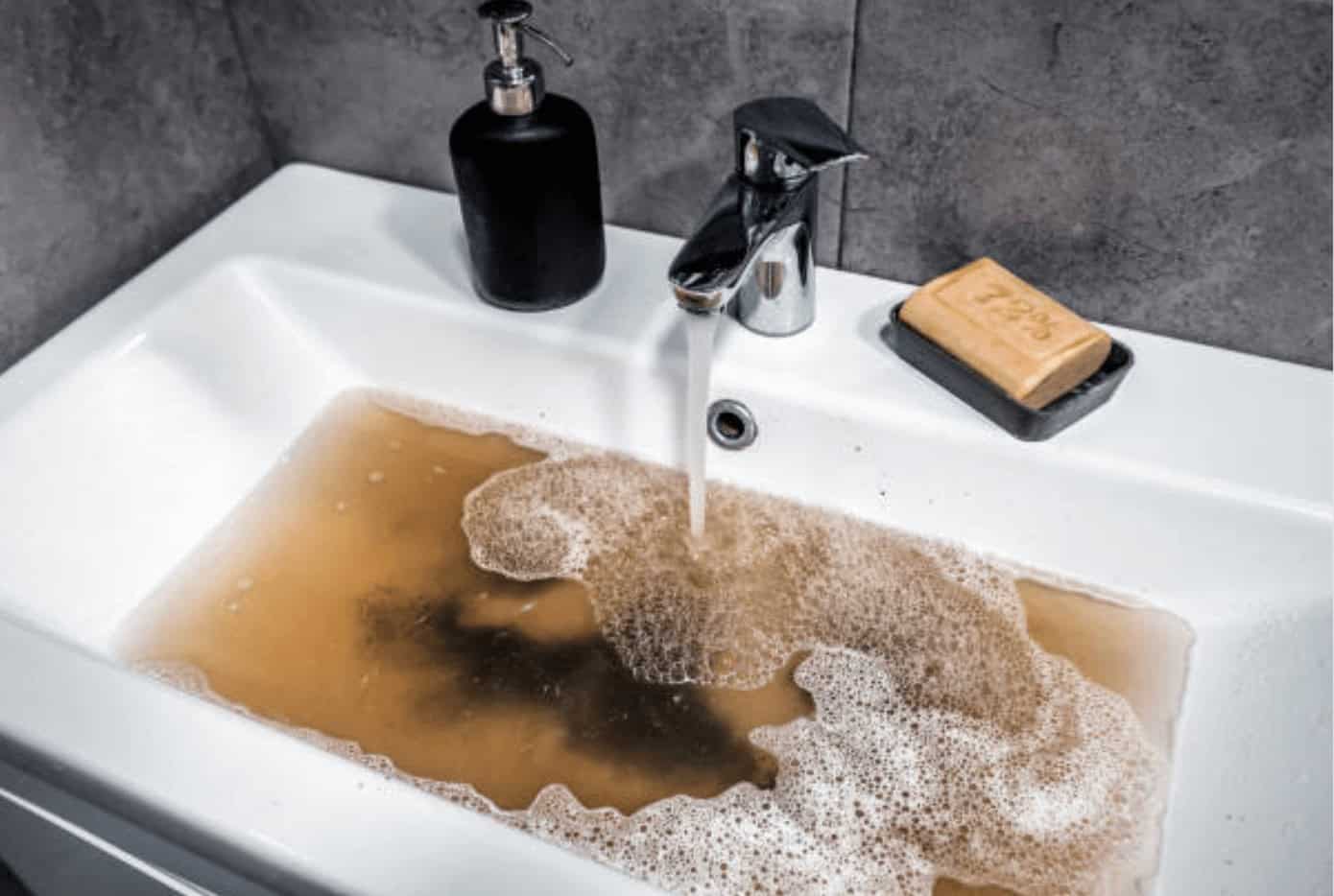 A white bathroom sink with murky brown water filling from the faucet, indicating a potential basement floor drain backup; Pipeworks Plumbing and Construction can resolve water quality and drainage issues. Soap and black dispenser on sink, gray tiled walls in background.