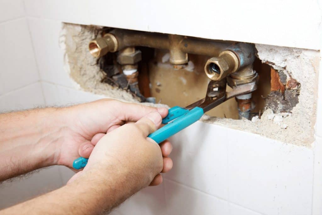 A Pipeworks Plumbing and Construction technician uses blue-handled pliers to repair exposed copper pipes inside a tiled wall, performing professional plumbing maintenance.