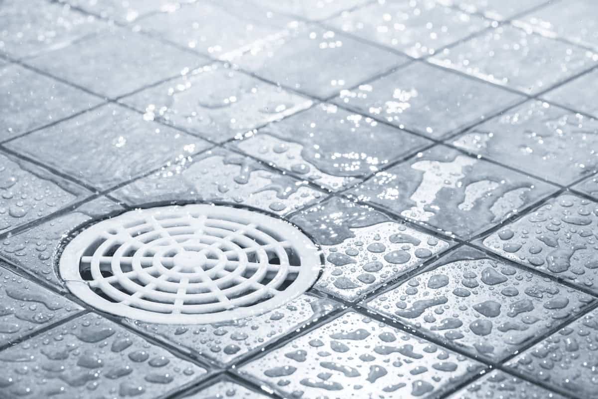 Wet, newly tiled bathroom floor with visible water droplets and a white circular drain cover, indicating recent pipe installation or repair by Pipeworks Plumbing and Construction.