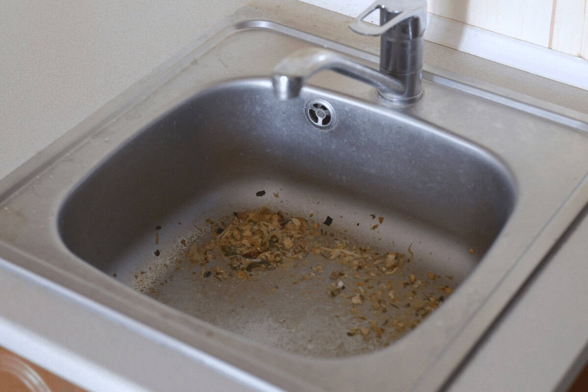 A dirty kitchen sink with food scraps and debris accumulating at the bottom, next to a chrome faucet—possible signs of a basement floor drain backup or clogged pipes; Pipeworks Plumbing and Construction can help resolve plumbing issues like this.