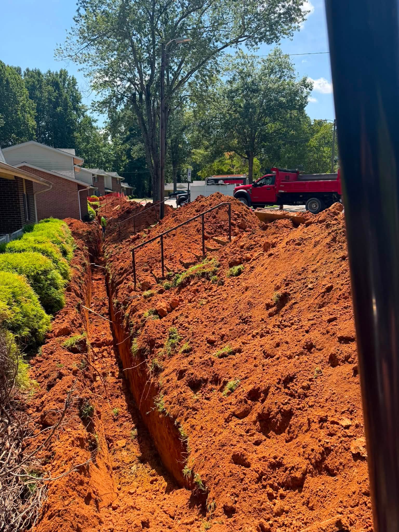 Deep water line trench running along occupied brick apartment buildings with handrails and a company dump truck staged for spoil haul-off