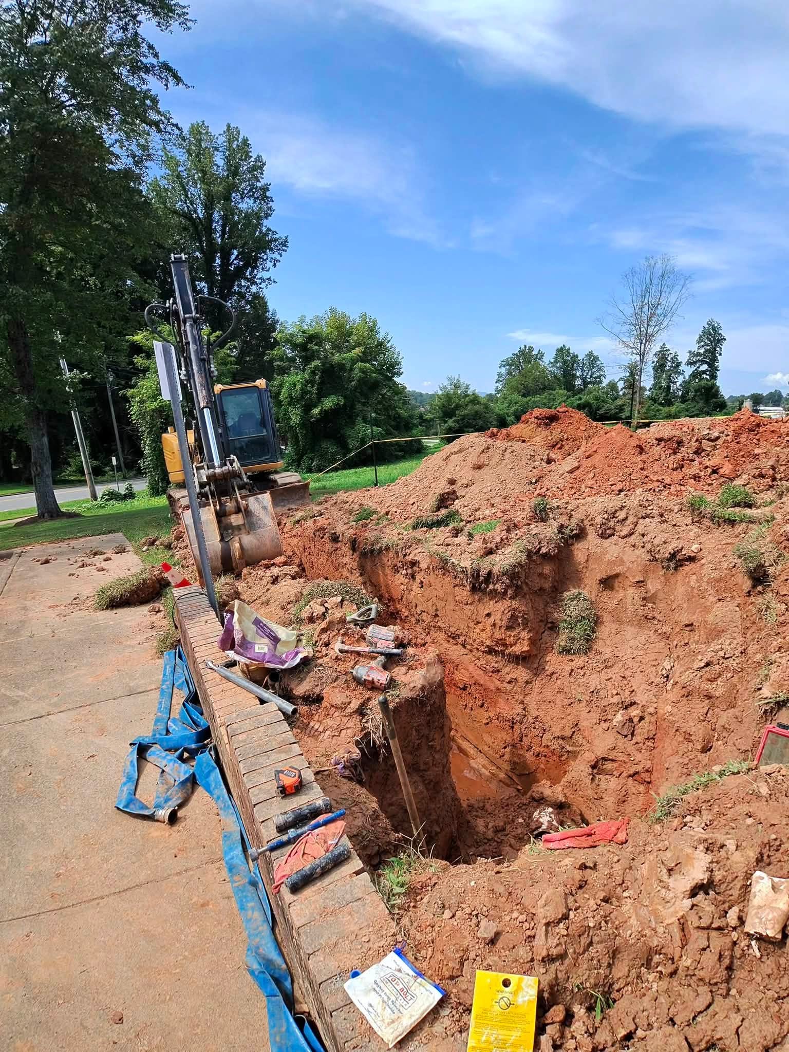 Tracked excavator working a deep water line connection pit beside a brick retaining wall with protective poly sheeting and caution tape