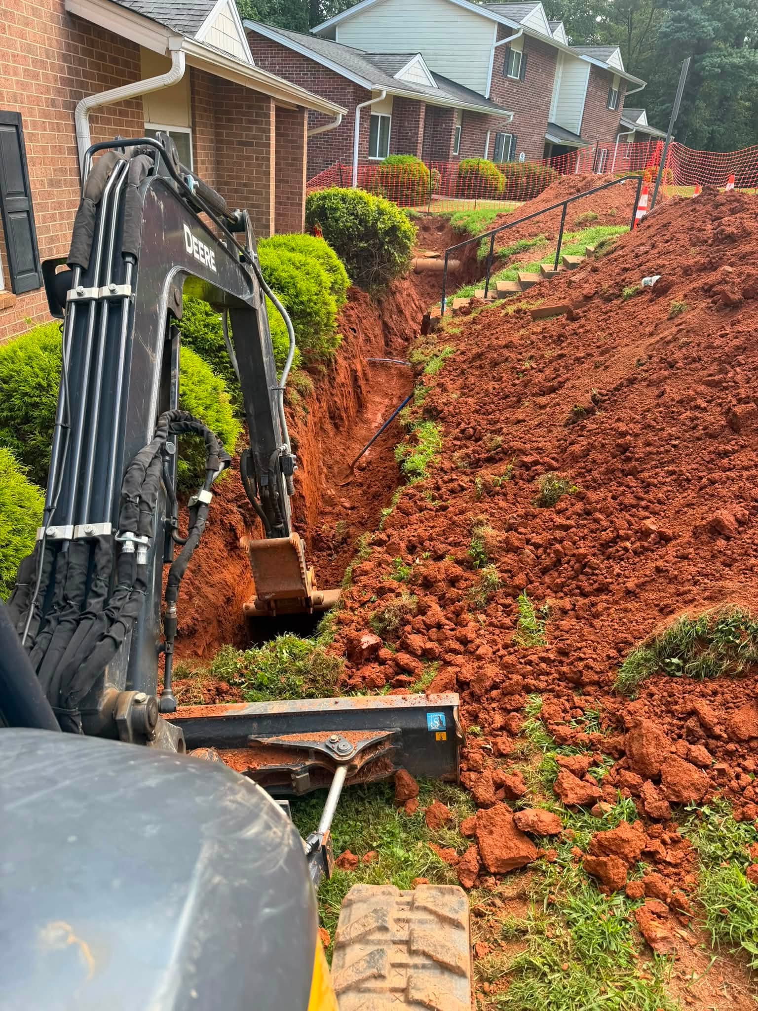 Mini excavator cutting a water line trench on a graded slope between an apartment foundation and mature shrub beds
