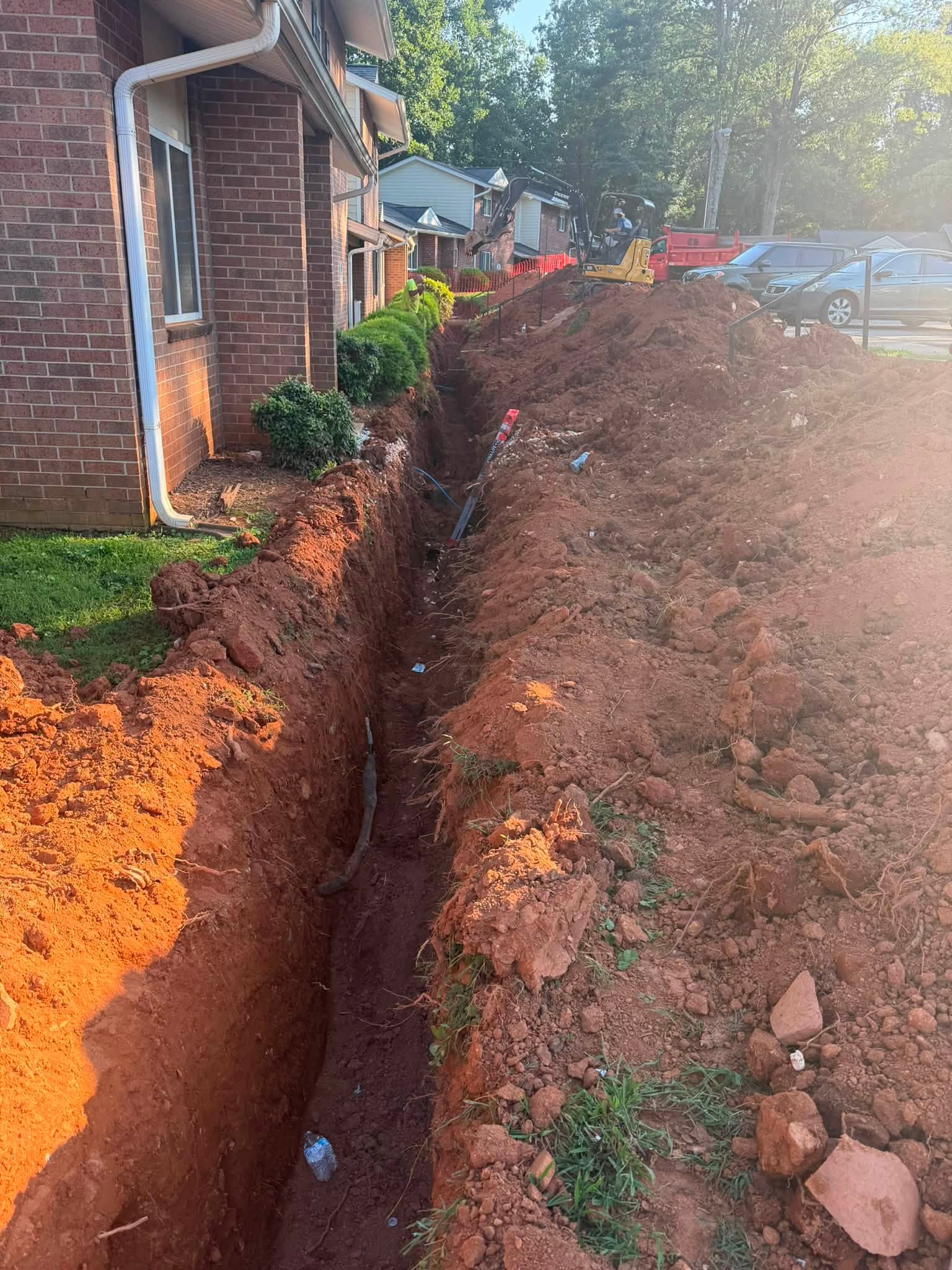 Narrow water service line trench beside an occupied building with the new pipe visible in the trench bottom