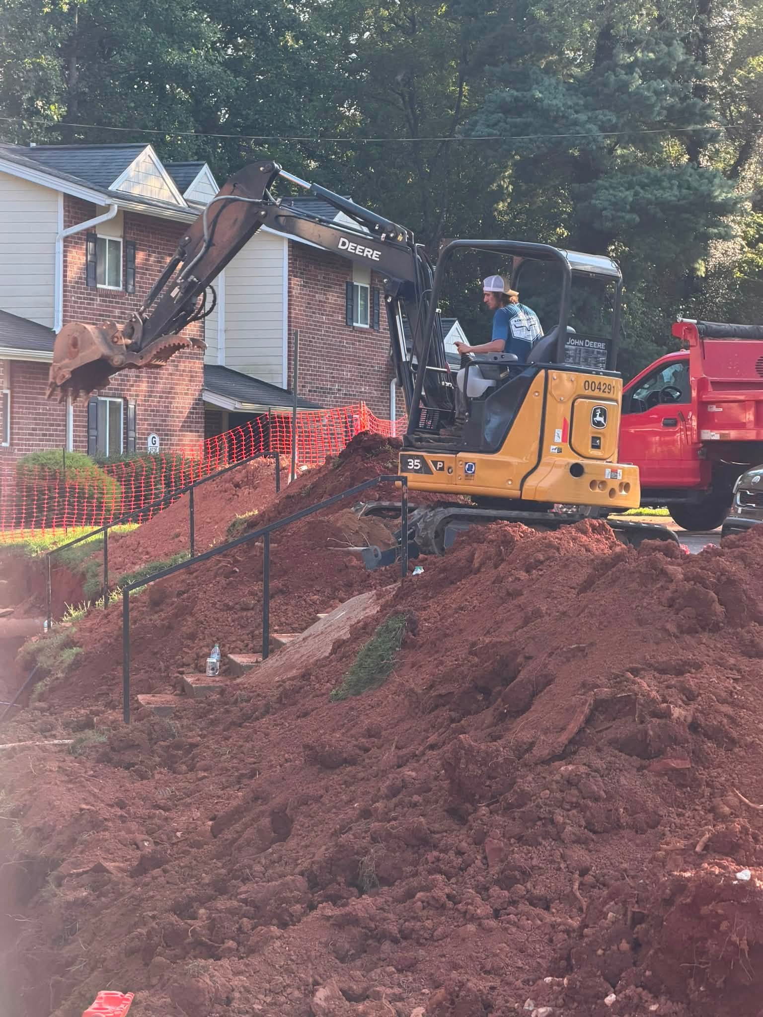 Pipeworks operator running a compact excavator beside safety fencing and a company dump truck at a multi-family water line replacement