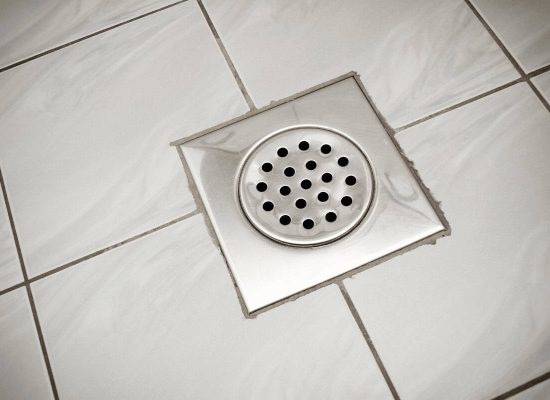 Close-up of a metal shower drain cover with round holes, installed in the center of square, light-colored bathroom tiles arranged in a grid pattern. The drain, professionally fitted by Pipeworks Plumbing and Construction, helps prevent basement floor drain backups and keeps your space dry. Close-up of a metal shower drain cover with round holes, installed in the center of square, light-colored bathroom tiles arranged in a grid pattern. The drain, professionally fitted by Pipeworks Plumbing and Construction, helps prevent basement floor drain backups and keeps your space dry.