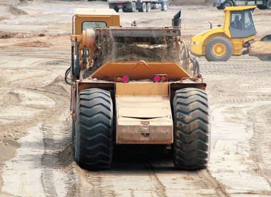 Pipeworks Plumbing and Construction earthmoving vehicle transporting soil at a commercial construction site for plumbing and sewer line installation, with additional excavators and trucks in the background. Pipeworks Plumbing and Construction earthmoving vehicle transporting soil at a commercial construction site for plumbing and sewer line installation, with additional excavators and trucks in the background.