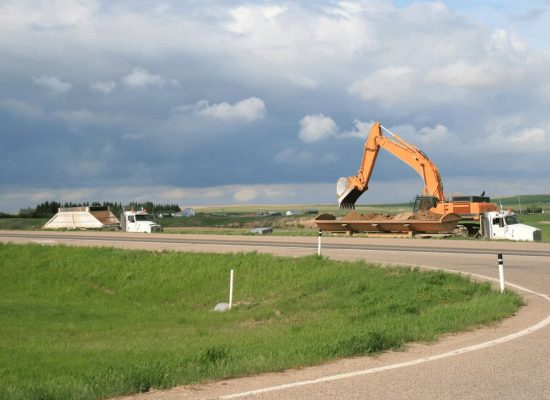 Pipeworks Plumbing and Construction orange excavator loading soil into a dump truck beside a rural highway, with green fields and distant trees under a cloudy sky; construction site for underground plumbing installation. Pipeworks Plumbing and Construction orange excavator loading soil into a dump truck beside a rural highway, with green fields and distant trees under a cloudy sky; construction site for underground plumbing installation.