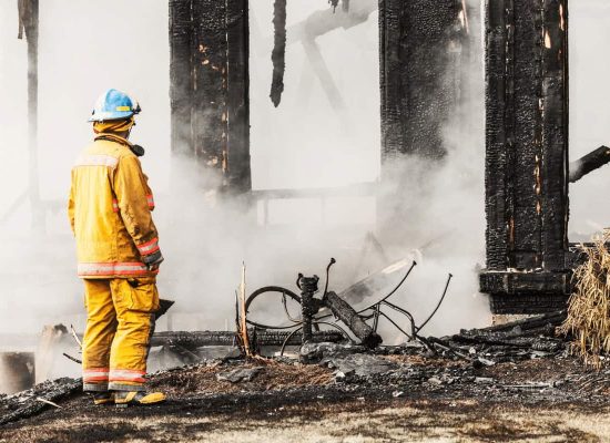 A firefighter in yellow gear and blue helmet surveys a fire-damaged building with charred debris and smoke, highlighting the consequences of basement floor drain backup; Pipeworks Plumbing and Construction specializes in preventing plumbing-related fire damage. A firefighter in yellow gear and blue helmet surveys a fire-damaged building with charred debris and smoke, highlighting the consequences of basement floor drain backup; Pipeworks Plumbing and Construction specializes in preventing plumbing-related fire damage.