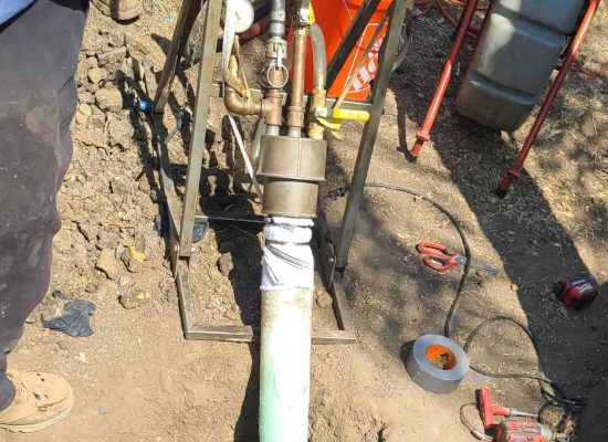Close-up of an outdoor pipe repair by Pipeworks Plumbing and Construction, showing plumbing tools, a hose attached to a pipe, an orange bucket, duct tape, and soil; two technicians legs are partially visible nearby. Close-up of an outdoor pipe repair by Pipeworks Plumbing and Construction, showing plumbing tools, a hose attached to a pipe, an orange bucket, duct tape, and soil; two technicians legs are partially visible nearby.
