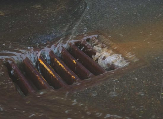 Rainwater flows into a metal storm drain on wet, dark pavement illuminated by yellow-orange streetlights, highlighting the crucial underground plumbing infrastructure managed by Pipeworks Plumbing and Construction. Rainwater flows into a metal storm drain on wet, dark pavement illuminated by yellow-orange streetlights, highlighting the crucial underground plumbing infrastructure managed by Pipeworks Plumbing and Construction.