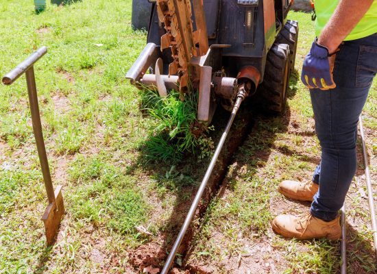 A Pipeworks Plumbing and Construction worker in gloves and work boots operates a trenching machine, digging a narrow trench in grassy soil for underground pipe installation, with plumbing tools and equipment nearby. A Pipeworks Plumbing and Construction worker in gloves and work boots operates a trenching machine, digging a narrow trench in grassy soil for underground pipe installation, with plumbing tools and equipment nearby.