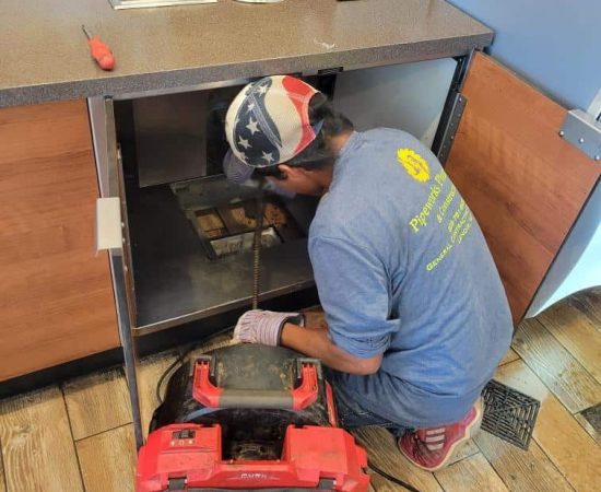 A Pipeworks Plumbing and Construction technician in a blue shirt and star-spangled cap kneels under a fast-food restaurant counter, using professional plumbing tools and a red drain cleaning machine to repair pipes after a basement floor drain backup.