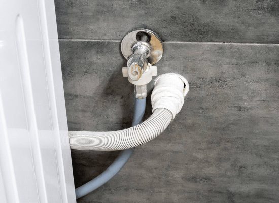 Close-up of washing machine water supply and drain hoses securely connected to wall-mounted valves and a drain pipe installed by Pipeworks Plumbing and Construction, set against a gray tiled wall; partial washing machine visible on the left. Close-up of washing machine water supply and drain hoses securely connected to wall-mounted valves and a drain pipe installed by Pipeworks Plumbing and Construction, set against a gray tiled wall; partial washing machine visible on the left.