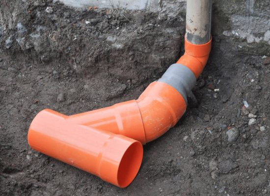 Partially buried orange PVC drainage pipe system installed by Pipeworks Plumbing and Construction; features a vertical section connected to a horizontal Y-fitting beside a concrete wall, illustrating proper basement plumbing and flood prevention. Partially buried orange PVC drainage pipe system installed by Pipeworks Plumbing and Construction; features a vertical section connected to a horizontal Y-fitting beside a concrete wall, illustrating proper basement plumbing and flood prevention.