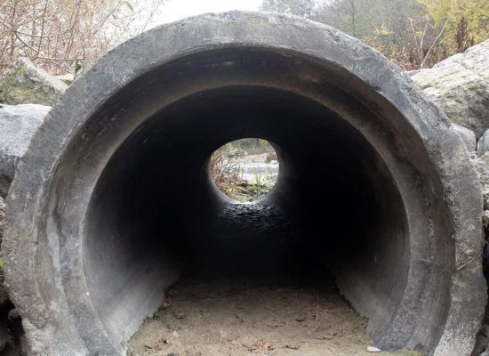 Large concrete drainage pipe installed by Pipeworks Plumbing and Construction, surrounded by rocks, with an open end revealing outdoor foliage—showcasing professional drainage installation in the construction industry. Large concrete drainage pipe installed by Pipeworks Plumbing and Construction, surrounded by rocks, with an open end revealing outdoor foliage—showcasing professional drainage installation in the construction industry.