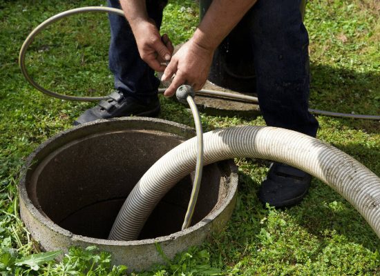 A Pipeworks Plumbing and Construction technician using a hose to pump and clean an outdoor septic tank, hands guiding a thin hose into the open tank on grassy ground—professional septic maintenance to prevent basement drain backups. A Pipeworks Plumbing and Construction technician using a hose to pump and clean an outdoor septic tank, hands guiding a thin hose into the open tank on grassy ground—professional septic maintenance to prevent basement drain backups.