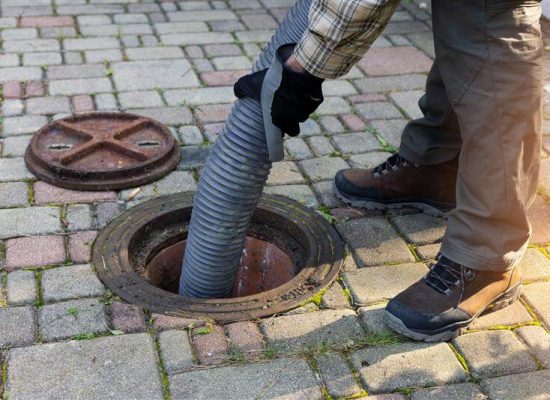 A Pipeworks Plumbing and Construction technician wearing gloves and work boots inserts a large industrial hose into an open manhole on a paved street, with the manhole cover removed—demonstrating professional sewer cleaning services. A Pipeworks Plumbing and Construction technician wearing gloves and work boots inserts a large industrial hose into an open manhole on a paved street, with the manhole cover removed—demonstrating professional sewer cleaning services.