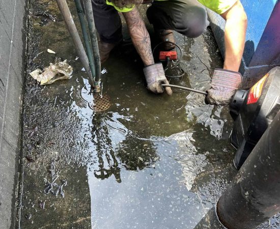 Pipeworks Plumbing and Construction technician in a neon yellow safety shirt kneels on wet basement floor, using plumbing tools to clear a clogged floor drain surrounded by standing water, debris, exposed pipes, and nearby blue construction equipment.